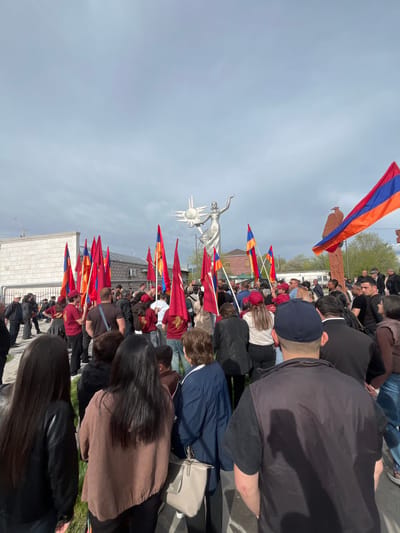 A crowd of people stand near the Mother Armenia statue in Akhuryan, Armenia. Many people are holding Armenian flags. The sky is overcast. Some buildings are visible in the background.