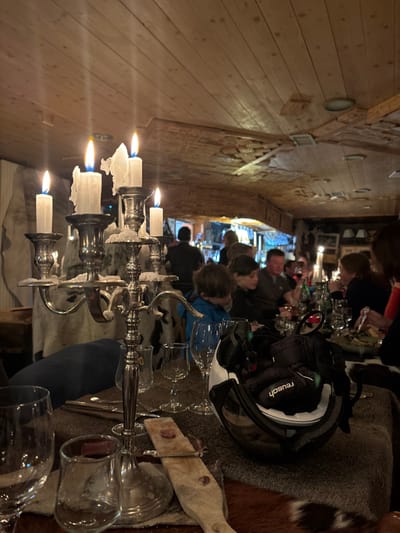 The image captures an indoor dinner scene, likely in the evening, with several people gathered around a long table in what appears to be a rustic restaurant or lodge. The overall lighting is dim and warm, primarily emanating from lit candles.
In the foreground, a prominent silver candelabra with several white candles is centered on the table. Many of the candles have melted wax dripping down, indicating they have been burning for some time. Glasses of various sizes are scattered across the table, some appearing to be wine glasses. A wooden serving board with a slice of cured meat or cheese is visible on the right. The table is covered with dark, furry textiles, possibly animal hides or faux fur, adding to the rustic ambiance.
Several individuals, including adults and at least one child (wearing a blue top), are seated along the table. Most are seen from behind or profile, engaged in conversation or eating. Their postures suggest a relaxed and social atmosphere. On the table, near the people, a black ski helmet with a white rim and clear visor is placed. The brand name "Reusch" is clearly visible in white text on the side of the helmet.
The interior architecture features a ceiling and upper walls clad in light-colored wooden planks, while a lower section of the wall appears to be constructed from exposed brickwork. The style reinforces the alpine or lodge setting. Given the presence of the ski helmet and the provided context of Val-d'Isère, France, the scene strongly suggests a meal following a day of skiing or winter activities in a mountain resort.