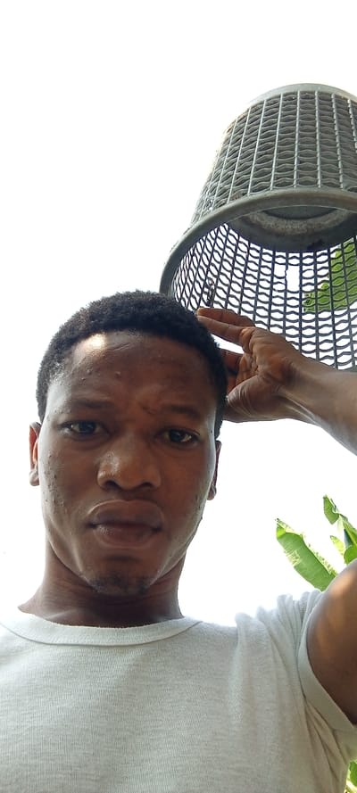 An outdoor, low-angle, head-and-shoulders portrait of an adult male captured in Aramoko-Ekiti, Nigeria. The individual, of African descent, features short, dark, curly hair and wears a light-colored, crew-neck t-shirt. He maintains a neutral expression, gazing directly at the camera. His right hand is raised, with fingertips touching the right temple region of his head. Dominating the upper right quadrant is an inverted, dark, perforated, basket-like object, from which a small metallic ring or hook hangs. Patches of green foliage are visible on the far right, set against a stark, brightly overexposed white sky.