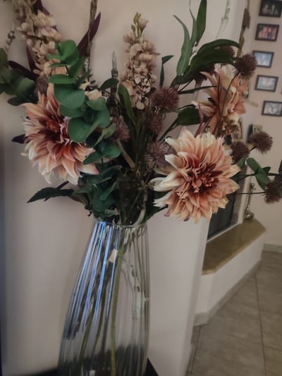 A vertical, slightly blurry, eye-level medium shot captures a bouquet of artificial flowers in a clear, ribbed glass vase. The flowers are a mix of peachy-pink dahlias and dried-looking elements in muted browns and purples, interspersed with green eucalyptus leaves. The vase is placed on a dark surface, with the lower half of the bouquet visible. The background consists of a light-colored wall and a section of a fireplace with a golden hearth and a tiled floor. Several small framed photographs are mounted on the wall in the upper right corner. The scene appears to be indoors, likely a domestic setting. The image lacks any human presence or activity, focusing solely on the floral arrangement and its immediate surroundings. No specific location in Trikala, Greece is identifiable from the visual elements.