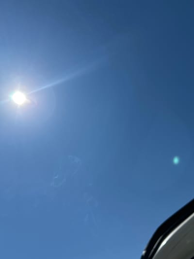 The image depicts a view of a clear, bright blue sky with the sun shining brightly at the bottom left corner. Faint wisps or trails appear to be present in the upper right section of the frame, possibly thin cirrus clouds or contrails. The top right corner shows a portion of an object, potentially a car frame or another vehicle component. It is impossible to confirm that this image was taken in the city of Mineralni Bani, Bulgaria, without additional information.