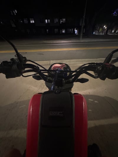 The image presents a first-person perspective from a stationary red motorcycle or scooter during nighttime in Gainesville, Florida, United States of America.

In the immediate foreground, the black handlebars, control levers, and visible cables of the motorcycle are centered. Attached to the handlebars, a blue lanyard with white text, partially readable as "UNIVERSITY," hangs down. The illuminated instrument cluster, displaying a reddish-orange glow, is visible below the handlebars. The central body of the motorcycle is bright red, featuring a black, textured, carbon-fiber-like panel. This panel includes a white rectangular label with indistinct text, likely a warning or instructional sticker. The rider's leg or foot is partially visible in the bottom left corner, suggesting an individual is seated on the vehicle.

Directly ahead, a two-lane asphalt road is visible, marked by double solid yellow lines indicating no passing. Beyond the road, a concrete curb or sidewalk separates the street from a row of dark, multi-story buildings. These buildings exhibit multiple illuminated windows, indicating internal occupancy or activated lighting. The architectural style suggests residential or commercial structures. Dark silhouettes of trees are discernible in the background, interspersed with the buildings, under the ambient glow of distant streetlights not fully within the frame. The overall lighting conditions are low, characteristic of an urban environment after dusk. No other individuals or vehicles are explicitly visible.