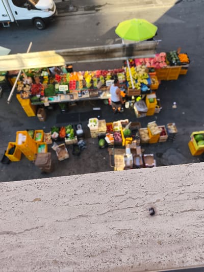 A street market is set up outdoors in Livorno, Italy, featuring stalls overflowing with fresh produce. A white van is parked to the left, partially visible. To the right of the van, a market stall displays a variety of fruits, including what appear to be watermelons, oranges, and bananas, arranged in crates. A bright green umbrella provides shade over another section of the market, where more fruits like apples, grapes, and possibly berries are displayed. In the foreground, a person wearing a white tank top and dark shorts is seen standing by a stall, interacting with the market setup. The ground is paved, and various empty and full crates are scattered around the market stalls. The scene suggests a vibrant commercial area within the city, likely a place where local vendors sell their goods to the public.