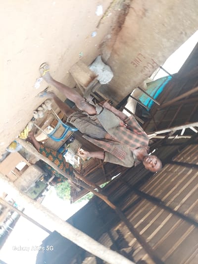 The image depicts an informal outdoor or semi-sheltered commercial area, assumed to be located in Awkuzu, Nigeria. The ground is exposed light brown soil with scattered debris, including paper. Overhead, a corrugated metal roof is visible on the right, supported by wooden beams and poles. A light blue tarp is present in the background to the right. A light-colored vertical surface, possibly a wall or board, displays the handwritten inscription "FOOD LINE."
Two adult individuals are present. In the mid-ground, a lean male with dark skin and short hair stands, facing slightly towards the camera. He wears a plaid long-sleeved shirt (red, black, and light tones), dark shorts, and two-toned sandals (one with a yellow strap, the other with a blue strap). His right hand holds a small, light-colored object, consistent with food. Near his feet are a crude wooden stool and a white sack on the ground.
To the left, an adult female with dark skin is positioned with her back mostly to the camera, attending to a counter or table surface. She wears a patterned dress or wrapper (green, brown, and orange motifs) and yellow-strapped sandals. On the surface in front of her are multiple small, spherical, light-colored items, consistent with produce, and a blue plastic container. The scene suggests daily commercial activity in a rustic environment.