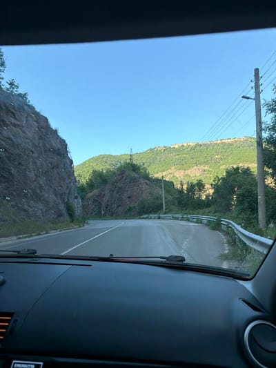 View from inside a vehicle traveling along a paved road in a mountainous region. On the left, a steep, rocky slope with wire mesh and vegetation is visible. Further up, a forested hillside stretches toward a clear blue sky with power lines. On the right, a metal guardrail lines the road. The car’s dashboard and windshield frame the scene. The location is Zasele, Bulgaria.
