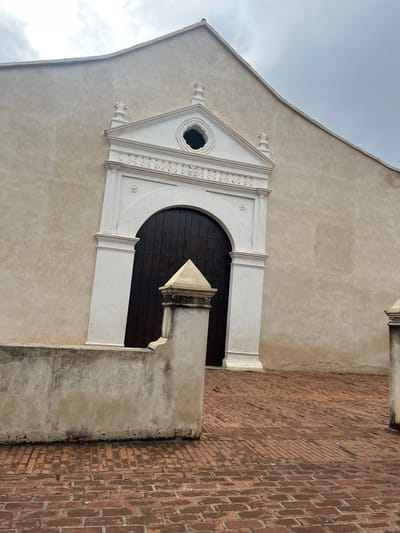 The image shows a close-up view of the facade of a building, likely a church or a historic structure, in La Asunción, Venezuela. The building has a light beige stucco exterior with a prominent white architectural embellishment above a dark wooden arched doorway. This embellishment includes decorative moldings, a triangular pediment with finials, and a circular window with intricate detailing.
In the foreground, a low, weathered wall made of concrete or stone runs horizontally across the lower part of the frame, partially obscuring a decorative stone pillar. The ground is paved with red bricks, some of which appear wet, suggesting recent rain or a damp environment. The sky above is overcast with gray clouds. The overall impression is one of historical architecture and a slightly somber atmosphere due to the cloudy weather.