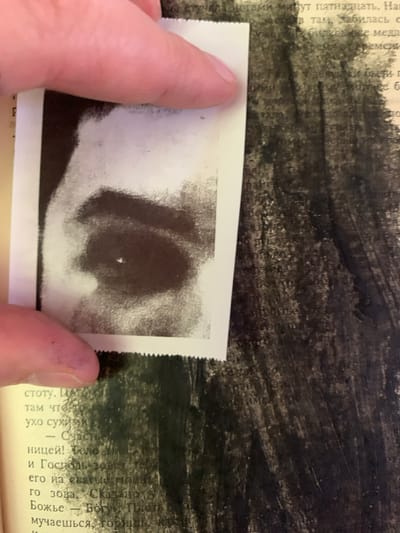 The image shows a close-up of a hand holding a black and white photograph over a textured, dark background that appears to be a page from a book or a canvas with brushstrokes. The photograph, partially obscured by the hand, is a close-up of what looks like an eye or a dark, amorphous shape with a bright speck in the center. The background is a dark, abstract pattern, possibly paint or ink, with some lighter areas that suggest texture or perhaps faint writing.

The text visible on the book page, in Russian, mentions "пятнадцать" (fifteen), "счастье" (happiness), "Господь" (Lord), and "Божье" (God's). The context of the text suggests a narrative or reflective content, possibly religious or philosophical. The location context provided is Ganino, Russia. The overall mood is introspective, with the combination of the personal photograph, the textured background, and the evocative text.