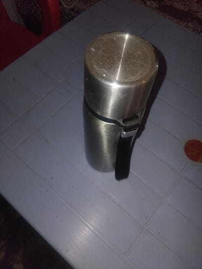 This media file shows a stainless steel thermos with a black handle resting on a light blue plastic surface. The thermos appears to be used, as there is some dust or residue on the top of it. The plastic surface has a grid pattern of indentations, suggesting it might be a table or tray. In the background, on the left side of the image, a patterned textile, possibly a rug or blanket, is visible. On the right side, a dark, out-of-focus area hints at the surrounding environment. There is also a circular object with red and orange colors visible on the plastic surface in the upper right portion of the image, which could be a label or a decorative element. No people are visible in the frame. The lighting appears to be artificial and somewhat dim, casting shadows. There is no indication of the time of day or weather. The overall scene is domestic or indoor.
