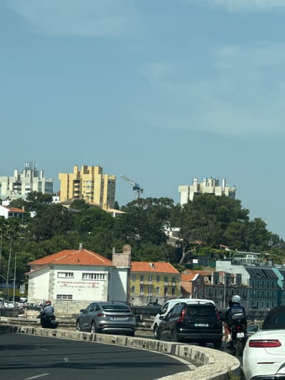 The image captures a scene on a sunny day in Oeiras, Portugal, likely along the Marginal road. In the foreground, a dark asphalt road curves to the left, with a concrete barrier separating it from a paved area. A silver Audi e-tron is parked on the paved area, facing left. To the left of the Audi, a person on a motorcycle wearing a blue shirt and red helmet is seen moving away from the camera. Further left, a few people are seated or lying on the ground near a white building with a red-tiled roof. This building has text that reads "AUTORIDADE MARÍTIMA NACIONAL" and "INSTITUTO DE SOCORROS A NÁUFRAGOS". Beyond this building, the landscape rises with lush green trees and several multi-story buildings, some with distinctive yellow and white facades. A construction crane is visible in the distance to the right. In the mid-ground and background, a mix of residential and commercial buildings are situated on a hillside, some exhibiting colorful facades. A white van and a black Dacia are also visible on the road. The sky is clear and blue.