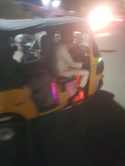 An overturned yellow vehicle, possibly an auto-rickshaw, is the central element. It is situated in an outdoor environment, likely at night, as suggested by the darkness. Light sources are visible, potentially streetlights and vehicle lights, causing significant lens flare. There appear to be people nearby, partially obscured and blurred due to motion or the lighting conditions. The location is possibly Dabawa, Nigeria.