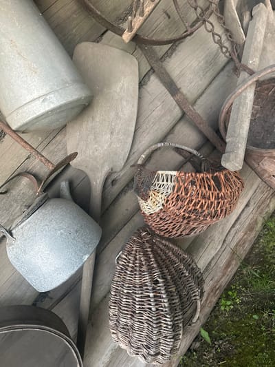 The image displays a collection of rustic, old farming or gardening tools and containers arranged on a weathered wooden deck. The subjects include two woven wicker baskets, one slightly broken, a large flat wooden paddle, two galvanized metal buckets or pots, and various rusted metal implements, including a chain and what appears to be part of a wheel assembly. The overall scene is one of aged, utilitarian items, suggesting a past use in a rural setting.
The objects are scattered, implying they are stored or perhaps discarded. There is no apparent activity taking place; the scene is static and observational. Notable details include the aged condition of the objects, with rust on the metal and wear on the wood and wicker, contributing to a sense of history and disuse. The wooden planks of the deck are also aged and weathered, with visible grain and gaps. The background shows a hint of green foliage and moss, suggesting an outdoor environment, possibly a garden or farmyard. The lighting appears to be natural daylight, likely overcast given the soft shadows.
There is no visible text in the image. The location context provided is Пярэжырскі сельскі Савет, Belarus, which aligns with the rustic and rural aesthetic of the objects. The overall mood is one of nostalgia and simplicity, evoking a connection to traditional rural life.
