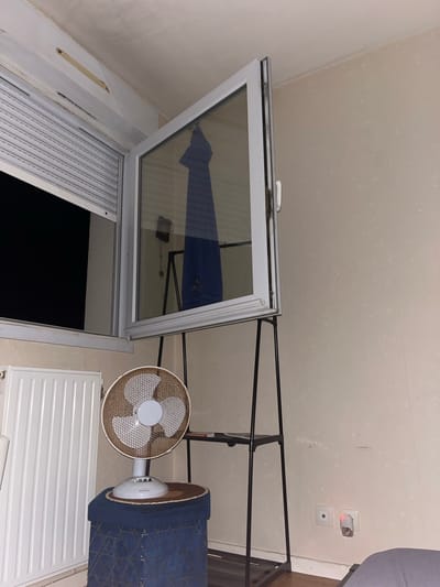 The image depicts an indoor scene, possibly a bedroom, featuring an open window with a roller shutter. A fan is placed on a patterned stool next to a white radiator. In the background, a black metal shelving unit stands against a textured wall. A small electrical outlet with a plug-in air freshener is visible on the wall. The overall impression is of a domestic environment, possibly in Chantepie, France, given the context provided.