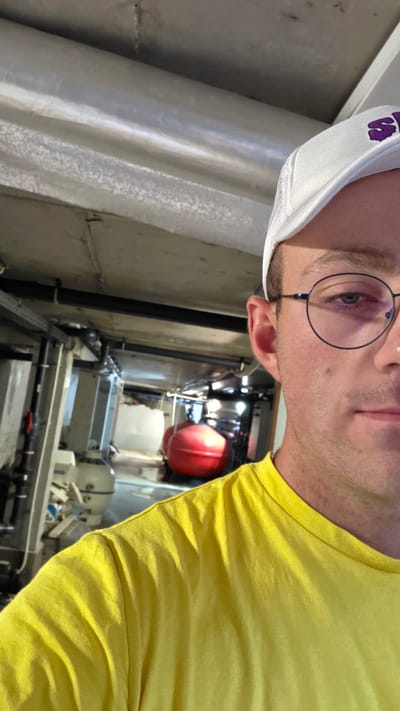 The image shows a close-up, waist-up, selfie-style shot of a man in a yellow t-shirt and a white cap with a purple logo. He is wearing glasses and has a slight stubble. In the background, there is industrial equipment, pipes, and a large red spherical object, possibly a tank or buoy. The environment appears to be an indoor industrial or utility space, possibly a facility or a boat. The lighting is artificial and appears somewhat dim. The image quality is moderate, with some graininess and visible compression artifacts. There is no discernible indication of the specific location in the city of Bania, Bulgaria.