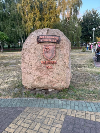 A large, rough, reddish-brown boulder stands in a grassy area, possibly a park or town square in Malaryta, Belarus. Attached to the boulder is a dark, metallic plaque bearing Cyrillic text and a coat of arms. The text on the plaque reads "ЛЮБЛЮ МОЙ КРАЙ, СТАРОНКУ ГЭТУ..." and below it "МАЛОРЫТА 1566". The coat of arms features a shield with a crown and what appears to be an oak leaf motif.
In the background, several tall, slender trees with lush green foliage dominate the scene. Sunlight filters through the leaves, casting dappled shadows. To the right of the boulder, a paved walkway is visible, made of interlocking bricks in shades of grey, yellow, and green. Several people are present in the background, some standing and talking near a lamppost and a fence. Others appear to be seated on benches. The overall impression is one of a public outdoor space, likely a memorial or historical marker, on a bright, sunny day.