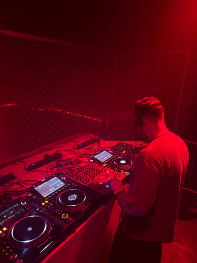 The image displays a DJ booth illuminated predominantly by an intense red light.
The image lacks specific visual identifiers (e.g., signage, architecture, landmarks) to confirm its location in Shanghai, China.
One individual is present, viewed from behind. This person appears male, has short dark hair, and is wearing over-ear headphones. He is dressed in a dark, long-sleeved shirt and dark trousers. His right hand is positioned on a control panel of the DJ mixer, suggesting active operation.
The DJ setup includes two Pioneer DJ media players flanking a central multi-channel DJ mixer, all resting on a light-colored countertop that reflects the red light. Both media players feature circular jog wheels and illuminated screens. The screen on the left player displays "DB" and timing information. The screen on the right player shows a waveform and "00:00:00.000" as the track time, along with "Pioneer DJ" branding on both units. The mixer is equipped with numerous knobs, faders, and illuminated buttons. A secondary arrangement of audio equipment is partially visible behind the main setup, further to the right.
Multiple audio and power cables are visible, connecting the equipment and draped across the counter. Two black power strips with blue indicator lights are positioned along the back edge of the counter.
The background features a dark, diamond-patterned chain-link fence or mesh barrier, stretching across the width of the scene and reflecting the red illumination. A dark, rectangular object, possibly an audio monitor, is situated behind the DJ, to the left of his head.
The scene's environment is characterized by the pervasive red light, which creates a specific ambiance and limits visibility of unlit details. The setting suggests an indoor venue, likely a club or event space.