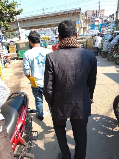 The image shows the backs of two men walking on a street next to a red motorcycle. The man in the foreground wears a dark suit and a checkered scarf. The other man has a light blue shirt with a star on the back and jeans with a yellow object in his pocket. Auto rickshaws are visible in the background, along with a concrete overpass and urban buildings under a bright blue sky.