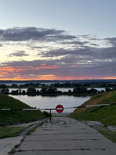 An image of a closed barrier across a concrete path leading down to a body of water, with a sunset in the background. The barrier is a horizontal bar with black and white stripes, and a red circular sign with a white horizontal line, indicating no entry. The concrete path is cracked and overgrown with grass. The body of water is calm and reflects the sky. The sky is filled with clouds, some of which are illuminated by the setting sun, creating a colorful display of oranges, pinks, and purples. The horizon is lined with trees and distant landforms. The location is identified as a barrier near the Volga River in the city of Bolgar, Russia.