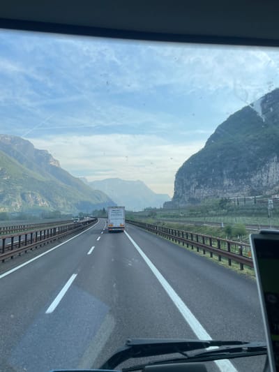 A medium-shot photograph taken from inside a vehicle shows a highway passing through a mountainous region, possibly in the city of Dolcè, Italy. The highway is flanked by guardrails and railway tracks on both sides. A white truck is traveling ahead in the same direction. The mountainous landscape is visible in the background under a partly cloudy sky. The image is taken through a windshield, with parts of the vehicle visible on the edges.