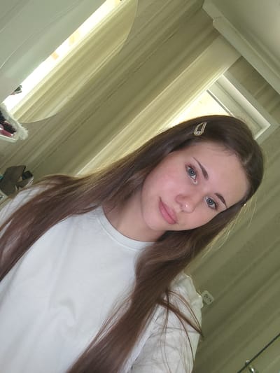 A young woman with long, brown hair is looking directly at the camera, taking a selfie. She is wearing a white t-shirt and has a sparkly hair clip in her hair. Her face is visible, with light makeup. She has blue-green eyes and pink lips. Her hair is long and straight, falling over her shoulders. The background appears to be a room with beige curtains and natural light coming from a window. There is a mirror in the background, reflecting the window and curtains. On a shelf or surface to the left, some personal items are visible, including what appears to be a hairbrush and some shoes. The overall setting is a domestic environment, possibly a bedroom. The city of Svietlahorsk, Belarus, is identified as the location of the image.