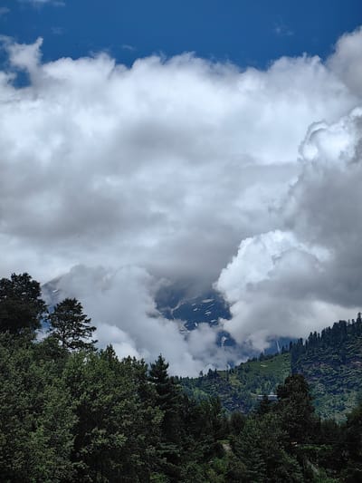 The image captures a vast, cloudy sky over a mountainous landscape with lush green trees in the foreground. The sky is a vibrant blue with large, fluffy white and gray clouds obscuring parts of the mountain peaks. A few small, dark specks, possibly birds or insects, are visible against the sky. The mountains themselves are rugged and covered in patches of snow, with dramatic cloud formations swirling around them. The foreground is dominated by dense, dark green trees, creating a sense of depth and framing the distant scenery. A hint of a building or structure can be seen on the mountainside, partially hidden by the trees and clouds. The overall impression is one of natural grandeur and a somewhat dramatic, overcast day in a mountainous region, potentially near Kulang, India, known for its scenic beauty.