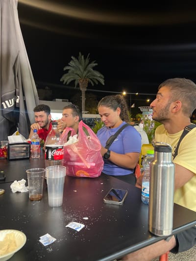 The image shows a group of four people seated at a black table outside a restaurant in Beersheba, Israel (31.251, 34.779). The people appear to be friends, enjoying a meal together. They are all dressed casually. Two men and two women are in the scene. One man is wearing a red shirt, another man is wearing a yellow shirt. One woman is wearing a blue shirt, and another woman is wearing a purple shirt. There is a large pink plastic bag on the table. One of the men is holding a bottle of Coca-Cola. One of the women is holding a clear plastic bag. A tall stainless steel water bottle sits on the table. In the background, there is a palm tree and the street. The street appears to be at night.  The scene is lit by streetlights. There are several empty glasses and plates on the table.