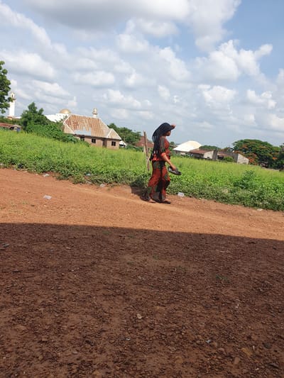 The image captures an outdoor scene in Idah, Nigeria, on a bright, cloudy day. A wide, reddish-brown dirt path or road dominates the right side of the frame, characterized by loose soil and small stones. To the left of the path, a strip of dense, vibrant green vegetation thrives, indicating a lush environment.
A person, likely a woman, is the central subject, positioned in the mid-ground on the edge of the dirt path. She is wearing a long, loose-fitting dress adorned with a complex pattern of red, orange, and black geometric and floral motifs, possibly an ankara fabric. Her head is covered with a plain black fabric, and she is wearing simple flip-flops. In her left hand, she carries a pair of dark-colored sandals. Her posture suggests she is either walking or pausing on her journey, looking towards the left side of the frame.
In the background, behind the green vegetation, various buildings are visible under the clear sky dotted with white cumulus clouds. Several structures feature sloped roofs made of corrugated tin. A notable architectural element is a building, possibly a mosque, which includes a distinct golden dome and one or more white minarets with conical tops, peeking above the green foliage. Trees with dark green leaves and some reddish flowers are also scattered in the distance. The lighting indicates it is daytime, likely late morning or early afternoon. No visible text is present in the image.