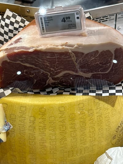 A close-up, eye-level shot focuses on a large piece of cured meat, possibly prosciutto, resting on a checkerboard-patterned paper lining. The meat exhibits distinct marbling and a thick layer of white fat. Attached to the meat is a small electronic price tag displaying "San Daniele Prosciutto Silver 1 kg" and a price of "4.99 /100g". Beneath the meat, a large wheel of aged cheese with a textured rind is visible. The cheese has embossed lettering, with "D.O.P." clearly legible in the lower portion of the frame. The background is blurred, but appears to be a retail environment, possibly a deli counter. This image was taken in Coquitlam, Canada, at a grocery store.