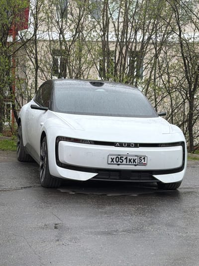 The image shows a white Audi e-tron GT parked on a paved surface, likely a street or parking area. The car's rear is facing the camera, revealing its taillights and the license plate "X051KK 51 RUS," indicating it is registered in Russia. The surrounding environment includes trees with budding leaves and a building in the background. Based on the license plate region code (51), which corresponds to the Murmansk Oblast, and given that Murmashi is the only city in the region, the photograph was taken in Murmashi, Russia.
