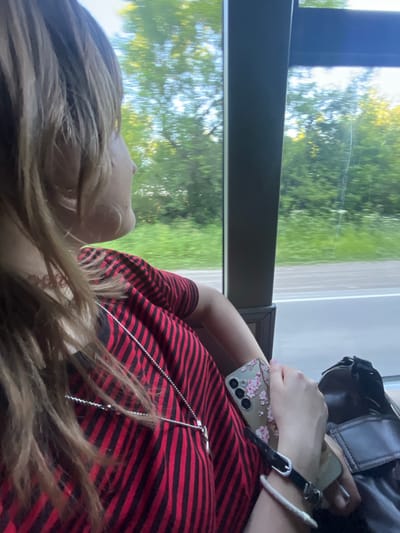 A person with light brown hair, wearing a red and black striped shirt and a silver necklace, sits next to a window, possibly on a bus or train. They are holding a smartphone with a floral case. The window shows a blurred view of trees and greenery, suggesting movement through a rural or park-like area, potentially on the outskirts of Kurtukovo, Russia. A dark bag is visible next to the person.