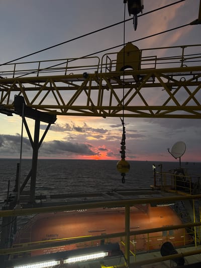 The image captures a dramatic sunset over the ocean, viewed from the deck of an offshore industrial platform. A large yellow crane structure dominates the foreground, with its intricate lattice work silhouetted against the colorful sky. A hook hangs from the crane's cable, suspended in mid-air. Below the crane, an orange lifeboat is visible, secured on the platform. The sea is calm, with a few distant vessels appearing as small specks on the horizon. The sky is filled with a palette of oranges, pinks, and grays from the setting sun, with scattered clouds adding texture to the scene. The overall impression is one of industrial might juxtaposed with the raw beauty of nature. The image location is not in Italy.