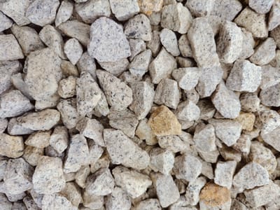 The image is an overhead close-up view of a pile of small, angular, light gray gravel with some flecks of black and occasional pieces of tan and light brown stone. The gravel is densely packed, creating a textured surface. There are no people, animals, or structures visible. The scene appears to be an outdoor environment, likely a ground surface or a pile of construction or landscaping material. Without further context, it is impossible to definitively determine the location within Palmiry, Poland.