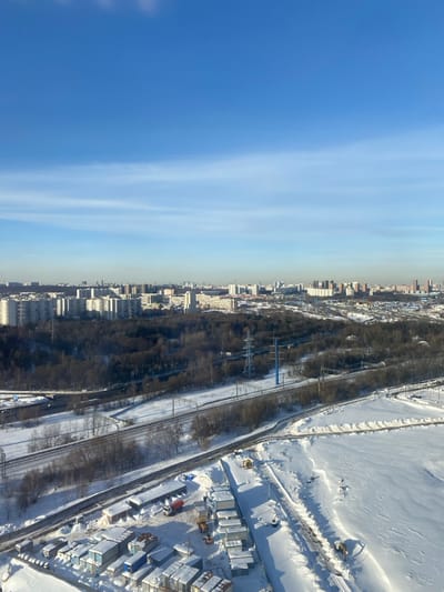 An aerial, high-angle view depicts a sprawling winter landscape in the city of Bittsa, Russia, under a clear blue sky with minimal cirrus clouds. The scene is dominated by snow-covered ground and urban infrastructure.
In the distant background, a dense urban skyline extends across the horizon, featuring numerous multi-story residential and commercial buildings. A visible haze lightly obscures the furthest structures.
The midground features a large, dark expanse of bare trees, indicative of a forested area or park. Running through this area are multiple parallel railway lines and associated roadways, all partially covered in snow. Several power line pylons are visible, including one distinct tall blue pylon.
The foreground presents a snow-covered industrial or construction site. Numerous modular container-like structures, some stacked, are arranged on the snow-covered ground. Visible vehicles include a red and white cement mixer truck and at least two yellow excavators or loaders. Cleared paths and excavated areas are present within the snowy terrain, suggesting ongoing activity. No individuals are discernible within the frame.