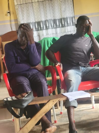 Two dark-skinned men are seated, one above the other, in red plastic chairs in what appears to be an interior space in Kwamba, Nigeria. The man on top wears a black shirt and jeans; he is touching his head with his right hand. The man below wears a purple long-sleeve shirt and pants and also has his hand on his head. A stack of papers rests on a wooden structure between the two chairs. The background includes patterned fabric, a green cloth, and a wall with a light-colored paint job. A pair of sandals are visible near the bottom.