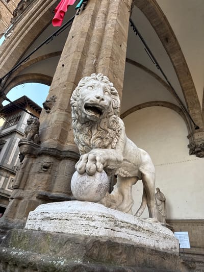 This image shows the "Marzocco" lion sculpture in Florence, Italy, located in front of the Palazzo Vecchio in Piazza della Signoria. The sculpture is made of marble and depicts a lion with an open mouth, paw resting on a sphere. The lion is positioned on a stone pedestal in the foreground. Behind the lion, a section of the Loggia dei Lanzi, an adjacent open-air gallery, is visible with its characteristic arches and stone columns. A red and green flag is hanging from one of the arches. Further in the background, a statue of a robed figure can be seen. The scene is lit by natural light, suggesting it was taken during the day.