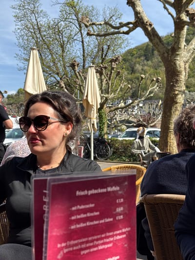 Here's a matter-of-fact description of the image:
**Scene:** An outdoor cafe setting.
**Location:** Cochem, Germany (based on the menu stating "OREL").
**People:** A woman with sunglasses is in the foreground, partially obscured by a red menu. Another person, potentially male, is seated across from her, partly visible at the top.
**Objects:**
* Menu with the title "Frisch gebackene Waffeln" (Freshly baked waffles) and listed prices.
* Sunglasses.
* Wicker chairs.
* Table umbrellas.
* Trees, including some with distinctive pruning.
* Buildings visible in the background.
* A bicycle.
* Vehicles, including what appears to be a white van.
**Action/Interaction:** Someone is looking at a menu.
**Environment:** The weather seems sunny. There's a view of buildings and greenery.