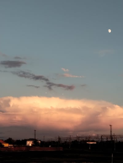 The image depicts a twilight scene in the city of Ақадыр, Kazakhstan. The sky is a gradient of pale blue and soft pink, with the moon visible as a small, bright orb in the upper right quadrant. Large, fluffy clouds dominate the middle ground, tinged with a peachy-pink hue from the setting sun. Below the clouds, a silhouetted landscape stretches across the horizon. This includes various structures, such as what appear to be railway tracks with overhead wires, utility poles, and some low-lying buildings or houses, one of which has illuminated windows. The foreground is a dark, indistinct area, possibly fields or undeveloped land. There are no people visible in the image.