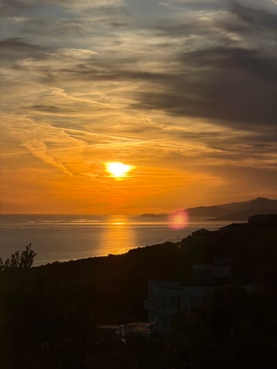 The image depicts a coastal sunset scene in Krimovice, Montenegro. The sun, a brilliant orange orb, is positioned in the upper center of the frame, casting a golden glow across the sky and the calm sea. Wispy clouds, painted in hues of orange, yellow, and gray, streak across the sky, creating a dynamic texture. The sea reflects the sunlight, forming a shimmering path of light that stretches towards the viewer.
In the foreground, a dark, silhouetted hillside slopes down towards the sea. Dotted along the hillside are several buildings, their structures mostly obscured by the deepening shadows. A prominent white, circular building with a distinct rooftop feature is visible in the lower right portion of the image. Trees and foliage are also visible in silhouette along the hillside. In the distance, the faint outlines of hills or mountains can be seen against the horizon. The overall atmosphere is serene and tranquil, characteristic of a sunset over the Adriatic Sea. No people are visible in the scene.