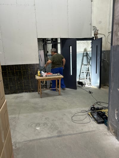 This media file depicts an indoor scene in Djerman, Bulgaria, likely a construction or renovation site. The image features a person wearing a green t-shirt and blue overalls, actively engaged in work. They are positioned near a wooden workbench, which holds various tools and supplies, including a yellow item that appears to be a drill, and what looks like a metal toolbox filled with fasteners.
In the background, a large, dark-colored door frame is being installed or worked on. A metal ladder or scaffolding structure is visible through the doorway, suggesting work at a higher level or within a confined space. The overall setting is industrial or unfinished, with concrete floors and unfinished walls covered in drywall. Cables and electrical equipment are present, indicating ongoing construction or electrical work.
The lighting suggests it is daytime, with artificial lights possibly supplemented by natural light from an unseen source. The atmosphere appears focused and industrious, with no clear indication of emotions beyond the concentration on the task at hand. The floor is marked with a patterned material, possibly insulation or protective sheeting, displaying the text "COSSMO" repeatedly. Other text on the wall panels is also visible, though partially obscured. The overall impression is that of a work in progress in a functional, non-domestic environment.