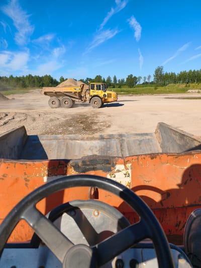 This is a first-person perspective photo taken from the driver's seat of what appears to be a large construction vehicle, possibly another dump truck or loader, looking out at a quarry or construction site. The foreground is dominated by the orange, weathered metal bed of the vehicle the photographer is in, and a black steering wheel.
In the mid-ground, a large yellow articulated dump truck is driving away from the viewer, kicking up dust. The truck is loaded with light-colored material, likely gravel or sand. It has "A25D" visible on its side. The ground of the site is dusty and sandy, with some tire tracks visible.
In the background, a line of green trees forms the horizon, with a clear blue sky above. Wispy white clouds are scattered across the sky, suggesting it's a sunny day, likely late morning or early afternoon given the bright sunlight and distinct shadows.
The overall impression is one of an active worksite on a clear, sunny day. There are no people visible, but the presence of heavy machinery and the dust being kicked up indicate ongoing operations. There is some faded text and warning labels on the orange vehicle in the foreground, but it is too blurry to read. The location is contextually identified as Skrunda-1, Latvia.