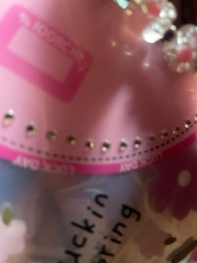 The image shows a close-up of pink and purple decorative items. On the left, a pink surface has what appears to be a translucent pink rectangle with a lighter pink outline and some text. Above this, there are blurred floral-shaped decorations with pink centers and clear, possibly beaded, petals. To the right of the pink surface, there is a lighter colored object with blurry floral patterns and some black text. The visible text on this object appears to read "buckin string" and "LUCKY DAY". The overall impression is of a collection of feminine, possibly craft or party supplies. The lighting is somewhat soft and diffused. The location context of Malete, Nigeria is noted, but there are no specific visual cues within the image to confirm this.