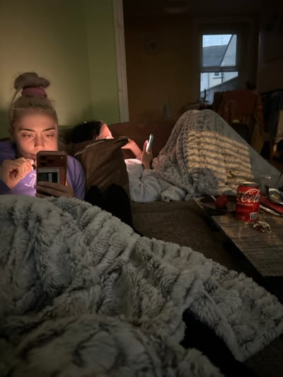 The image depicts an indoor scene within a residential dwelling located in Dorchester, United Kingdom, under dim lighting conditions. Two individuals, both appearing to be female, are situated on a dark-colored sofa.
In the foreground, on the left, a young adult female with blonde hair tied in a messy bun with a pink scrunchie is visible. She wears a light purple long-sleeved top. Her left hand holds a dark-colored smartphone, illuminated by its screen, which casts a glow on her face. Her right hand is near her mouth, holding a small, white, cylindrical object. A light grey and white textured blanket covers her lap and extends into the lower foreground.
Behind and to the right of the first individual, partially obscured, a second person is reclined on the sofa, facing away from the camera. This individual wears a light-colored long-sleeved top and holds a light-colored smartphone with their left hand, its screen also emitting light. They are covered by a light grey textured blanket, which displays distinct horizontal shadow stripes, indicative of light passing through window blinds. A large brown pillow is positioned between the two individuals.
To the right, on a dark-colored surface or table, a rectangular woven placemat holds a red Coca-Cola can, a small red object (possibly a lollipop), and a small, shiny metallic object.
The background reveals a room with a light green wall on the left and a warm brownish-tan wall on the right. A window with horizontal blinds is visible on the right, showing an exterior view of residential rooftops and part of a building. The overall lighting is low, with smartphone screens serving as primary light sources in the immediate vicinity of the individuals.
