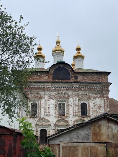 The image shows a close-up, low-angle view of the facade of an old, white brick church with golden domes, located in the city of Кстинино, Russia. The church exhibits signs of age and disrepair, with peeling paint and weathered brickwork. Several golden domes, characteristic of Russian Orthodox churches, are visible at the top of the structure. In the foreground, a large, leafy green tree partially obstructs the view of the church. Below the church, a portion of a weathered, rust-colored metal structure and a roofed shed are visible, obscuring the lower part of the church's facade and base. The sky is overcast and a light gray. There are no people or animals visible in the image.