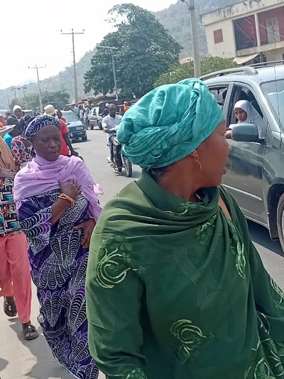 The image depicts a street scene in Ogaminan, Nigeria, under bright daylight conditions. In the foreground, a woman wearing a green headwrap and an embroidered green top or shawl is positioned on the right, facing right and partially obscured by the frame. She exhibits a gold earring. To her left, a woman in a light purple headwrap and a purple patterned wrapper with white and black designs walks forward. Her right wrist displays multiple gold bracelets. A third individual in pink pants and dark sandals is partially visible behind the woman in purple.
An open-windowed, dark-colored SUV occupies the mid-ground on the right, revealing a female occupant wearing a white hijab, looking left. On the left side of the road, a man operates a motorcycle, preceding other vehicles and pedestrians including a dark sedan. The environment includes multiple utility poles with overhead wires and streetlights. In the background, hills or mountains are visible, densely covered with vegetation including a prominent large tree. Several buildings are also present along the street.