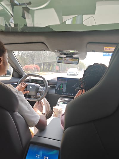 The image presents a forward-facing view from inside a modern vehicle, traveling in Jurong, China. An adult male, appearing to have dark skin and short dark hair, occupies the driver's seat, wearing a light-colored short-sleeved shirt and light-colored pants. His hands are positioned on the grey, multi-function steering wheel, which bears an "XPeng" logo. A digital display behind the steering wheel reads "52 D". To the right, the back of a second individual's head, with dark dreadlocks or braids, is partially visible in the front passenger seat.

The vehicle's interior features a large central infotainment screen displaying a navigation map interface with green route lines and media playback controls featuring Chinese characters. On the center console, a light-colored cup with cartoon-like figures rests in a cup holder. Below, an additional screen on the console displays "11 27". The car is equipped with a panoramic glass roof. A digital rearview mirror shows the road behind the vehicle.

Through the windshield, a paved road is visible under clear daylight. Other vehicles are present in the distance, including a white sedan directly ahead. The roadside environment features trees and bushes, with some foliage displaying reddish-orange hues. A blue and white road sign, partially obscured, with Chinese characters and a white cross symbol, is visible on the right side of the road, confirming the Chinese locale. The vehicle is in motion.