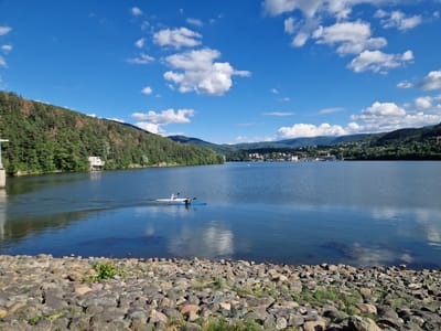 The image depicts a scenic view of Pancharevo Lake in Pancharevo, Bulgaria, under a clear blue sky dotted with white cumulus clouds. In the foreground, a rocky shoreline with scattered grass leads down to the water's edge. The lake itself is calm and reflects the sky and surrounding landscape.
On the water, two individuals are engaged in kayaking. One kayaker is in a white kayak, positioned slightly behind and to the left of another kayaker who is in a blue kayak. Both are paddling forward, leaving a wake on the water's surface.
The middle ground of the image shows a densely forested hillside on the left, with some buildings nestled amongst the trees. The far bank of the lake is lined with a town or village, characterized by numerous buildings and infrastructure, including what appears to be a harbor with several boats. The landscape beyond the town rises into rolling, tree-covered hills and mountains, disappearing into the distance. The overall impression is a tranquil natural setting complemented by human activity and settlement.