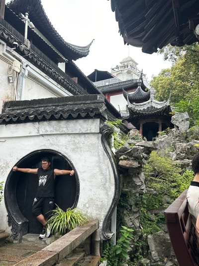 A man stands with his arms outstretched in a circular doorway of a traditional Chinese building in the Yu Garden, Shanghai, China. He is wearing a black sleeveless shirt with white lettering, black shorts, and athletic shoes. The building features dark, ornate roof tiles and white walls. To the right of the building, a rocky landscape with lush green foliage and a traditional Chinese pavilion is visible. In the background, a white building with a dome can be seen above the trees. A person's shoulder and back are visible in the lower right corner of the image.