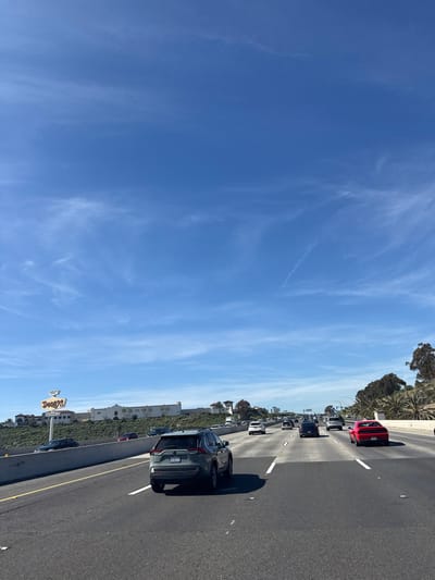A highway is depicted under a bright blue sky with wispy clouds. Multiple lanes of traffic are visible with various vehicles, including a red car, a dark SUV, and others further down the road. The shoulder of the highway shows some vegetation, a building, and a Denny's restaurant sign. The overall scene conveys a typical highway view on a clear day.
