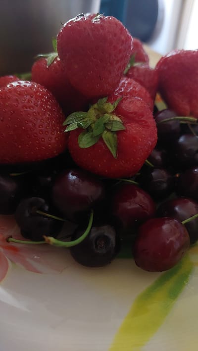 A close-up shot shows a pile of fresh strawberries and cherries in a bowl. The strawberries are bright red and plump, with green stems and leaves still attached. The cherries are a deep, dark red, almost black, with slender green stems. Some of the cherries have a glossy sheen, indicating they are ripe and juicy. The fruits are clustered together, creating a vibrant and appealing composition. The bowl itself appears to be made of ceramic and has a decorative pattern of yellow and green stripes on the rim, suggesting it's meant for serving. The lighting is bright, likely from natural daylight, illuminating the textures and colors of the fruit. The overall impression is one of freshness and abundance, perhaps a snapshot taken in a kitchen or during a meal preparation. The Lermontov, Russia location context doesn't offer specific visual cues within the image itself.