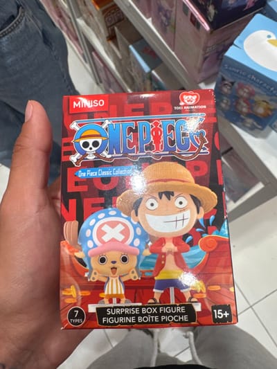 A human hand is holding a red MINISO-branded "One Piece Classic Collection" surprise box figure in a retail store environment. The box prominently features illustrations of the characters Monkey D. Luffy and Tony Tony Chopper. The text "SURPRISE BOX FIGURE," "FIGURINE BOITE PIOCHE," "7 TYPES," and "15+" is visible on the lower portion of the box. "TOEI ANIMATION" is also visible in the upper right. The background consists of white retail shelving displaying various other boxed merchandise, including a blue box with "Sanrio character" text and a cartoon duck design. A section of blue jeans and a white drawstring hoodie are visible at the bottom of the frame, indicating the presence of another individual or the lower body of the person holding the box. The scene takes place within a MINISO store located in León, Mexico.