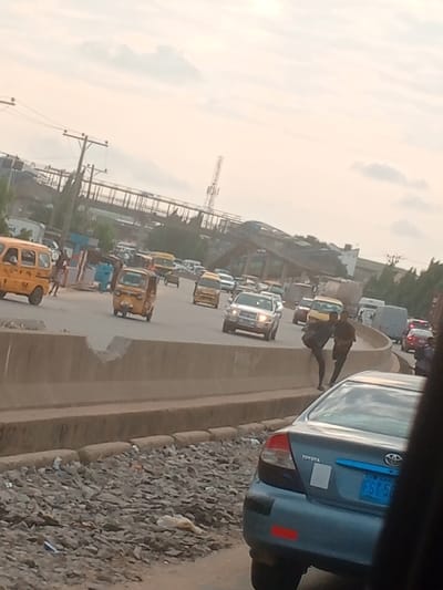 The image depicts a daytime urban street scene in Abule Egba, Nigeria, under an overcast sky. In the immediate foreground, the rear section of a light blue Toyota sedan, bearing a Nigerian license plate "FST 5E", is partially visible. Adjacent to the vehicle, a rough, unpaved area strewn with rubble and debris leads to a concrete median barrier. This barrier exhibits significant structural damage, including a large, irregular void.

Two dark-skinned males, dressed in dark clothing, are positioned on or in the immediate vicinity of this damaged barrier. One male is observed in a crouched, climbing, or descending posture, while the second male stands nearby. One of the males carries a bag or object slung over his shoulder.

Beyond the barrier, a multi-lane road extends into the background, accommodating moderate to heavy traffic, primarily moving away from the viewer. The vehicles include multiple yellow commercial minibuses, several yellow three-wheeled auto-rickshaws, various passenger cars, and SUVs, notably a silver SUV with illuminated headlights. In the far background, a white van is discernible. On the far left side of the road, several pedestrians are visible, with one individual prominently wearing a light blue garment. Overhead, an elevated pedestrian bridge or flyover structure spans the road. Electrical utility poles with overhead lines and a telecommunications mast are also present in the background.
